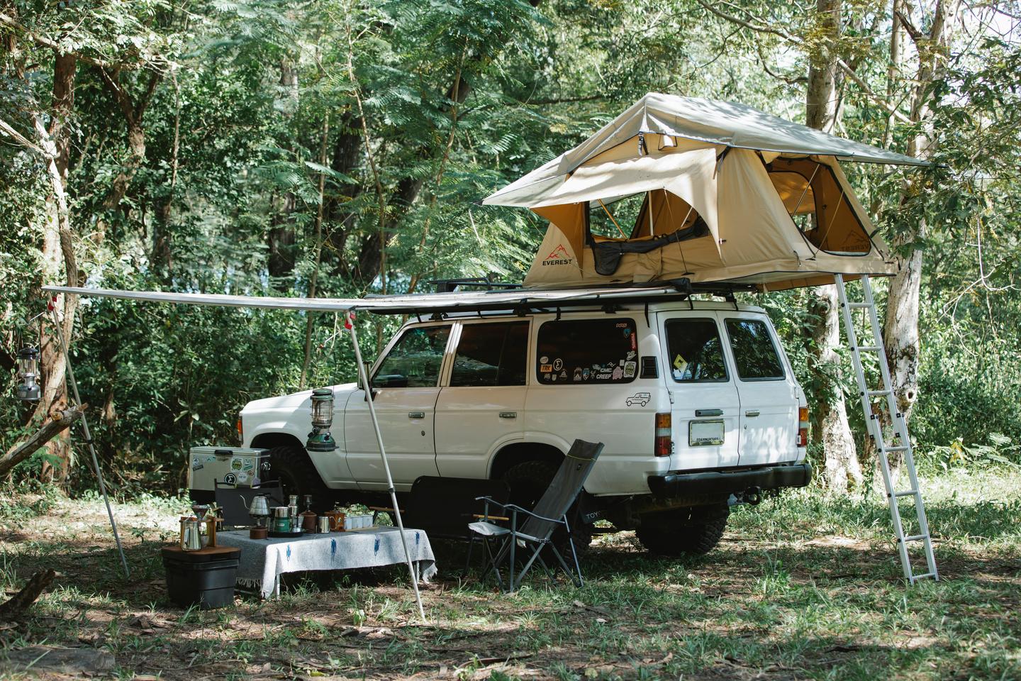 White off-road SUV with rooftop tent set up in a lush forest clearing, showcasing adventure vehicle preparation